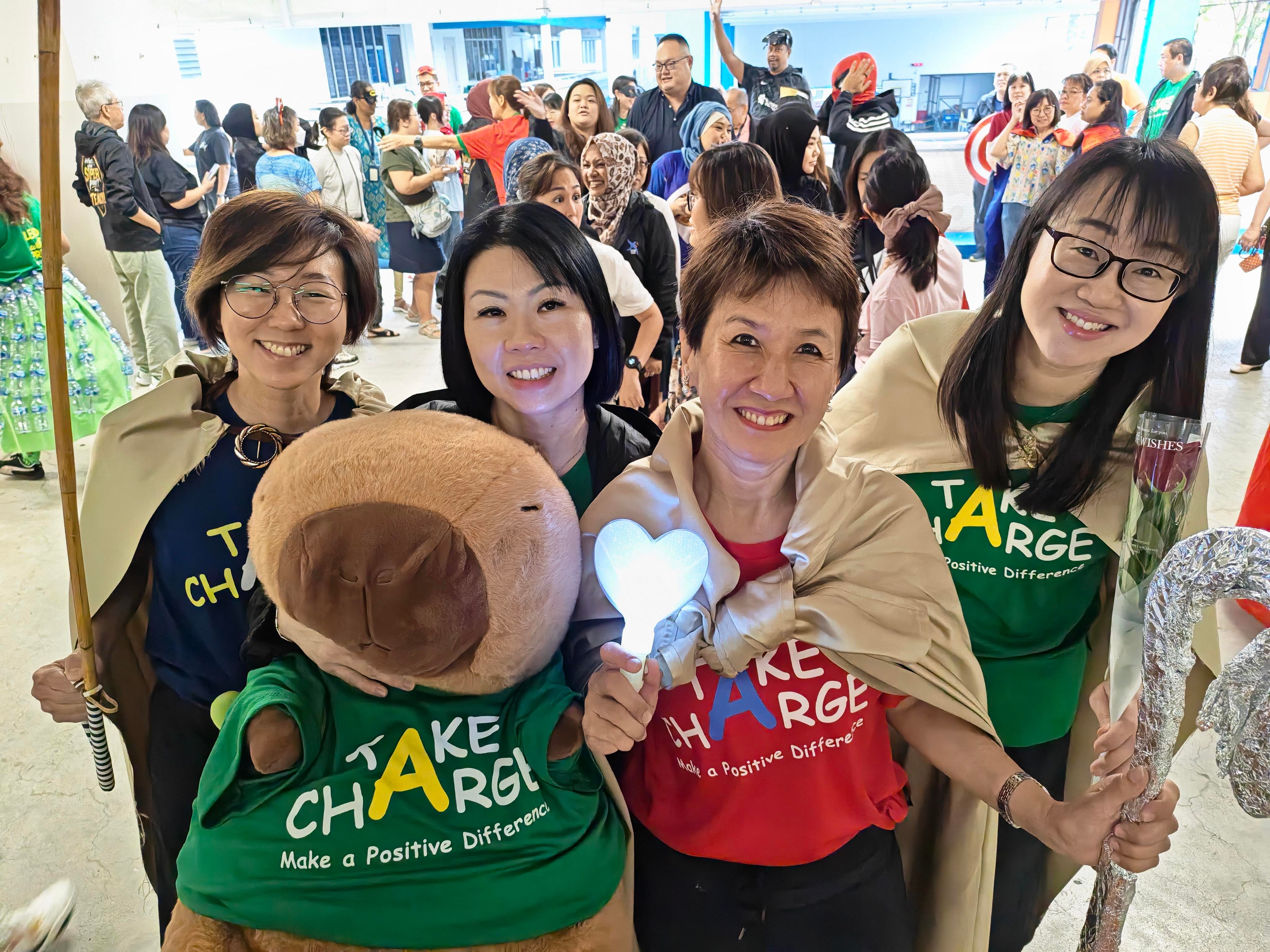 Four smiling school leaders pose with a large stuffed animal mascot wearing a "Take Charge, Make a Positive Difference" T-shirt, during the Teachers' Day red carpet walk-in. Staff and guests are gathered in the background.